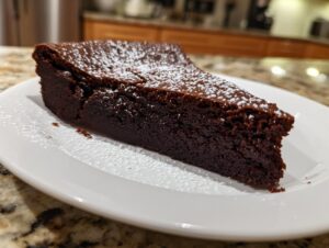 A close-up of a dense, dark slice of flourless chocolate cake dusted lightly with powdered sugar on a white plate.