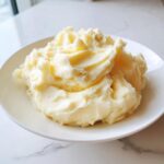 A generous serving of creamy, fluffy mashed potatoes piled high on a white plate.