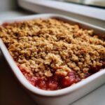 A close-up view of a freshly baked rhubarb crisp with a thick, golden, crumbly oat topping over bubbling fruit filling.
