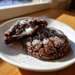 Two rich, dark hot chocolate cookies dusted with powdered sugar, one broken open to show gooey marshmallow and chocolate chips.