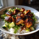 Close-up of a bowl filled with white rice, fresh greens, cucumber chunks, and pieces of glazed harissa chicken.