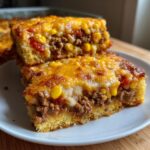 Two stacked squares of Mexican cornbread casserole featuring seasoned ground meat, corn, tomatoes, and melted cheddar cheese topping.