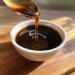 A spoonful of thick, glossy homemade teriyaki sauce is being poured into a small white bowl.