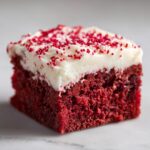 Close-up of a square slice of moist red velvet poke cake topped with thick white frosting and red sprinkles.