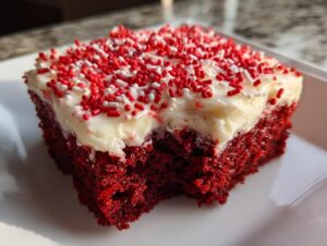 Close-up of a moist slice of red velvet poke cake topped with thick white frosting and red and white sprinkles.