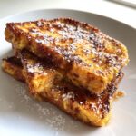 A stack of three slices of golden brown sourdough french toast dusted with powdered sugar and drizzled with syrup.