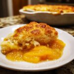 A close-up of a serving of southern peach cobbler with a golden crust and syrupy peaches on a white plate.