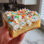 A close-up of a thick slice of sugar cookie bars topped with white frosting and colorful sprinkles.