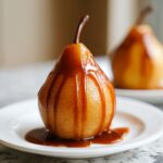 A beautifully presented poached pear drizzled generously with thick caramel sauce, sitting on a white plate.