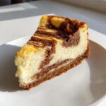 A close-up slice of marbled sugar cookie cheesecake with a graham cracker crust, sitting on a white plate in bright light.