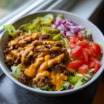A close-up of a tasty cheeseburger bowl featuring seasoned ground beef, melted cheese sauce, lettuce, tomatoes, pickles, and red onion.