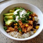 A vibrant, close-up view of a delicious chicken burrito bowl layered over white rice with black beans, corn, avocado slices, sour cream, and cilantro.