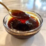 A close-up of thick, dark homemade teriyaki sauce being lifted from a small glass bowl with a golden spoon.