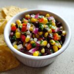 Close-up of colorful cowboy caviar featuring black beans, corn, red onion, and peppers, served in a white bowl next to tortilla chips.