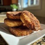 A stack of four round, thick churro cookies heavily coated in cinnamon sugar, resting on a white plate.