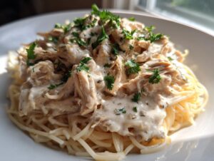 A close-up of shredded crock pot angel chicken in a creamy sauce served over a bed of spaghetti, garnished with parsley.