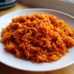 A close-up, appetizing view of vibrant, orange-red jollof rice served on a white plate.