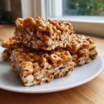 A stack of four gooey, golden-brown cheerio bars resting on a white plate near a window.
