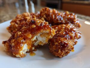 Close-up of crispy fried chicken tenders coated in a thick, glossy hot honey sauce, showing the white interior of one piece.