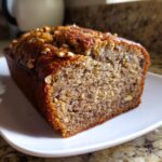 Close-up of a thick slice of moist protein banana bread topped with chopped walnuts, resting on a white plate.