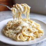 A fork lifts fettuccine noodles coated in creamy cottage cheese alfredo sauce, showing long, delicious cheese pulls.