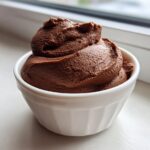 A serving of rich, dark chocolate mousse piped high in a small white ramekin, set against a bright window background.