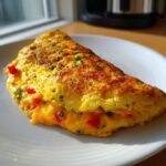A perfectly folded air fryer breakfast omelette filled with melted cheddar cheese and red peppers, sprinkled with herbs.