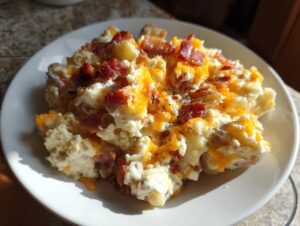 A serving of creamy baked potato salad topped generously with melted cheddar cheese and crispy bacon bits.