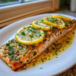 A close-up of a flaky baked salmon recipe fillet topped with lemon slices, garlic, and fresh parsley in a buttery sauce.