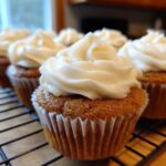 A close-up shot of freshly baked banana cupcakes topped with thick, creamy white frosting, cooling on a wire rack.