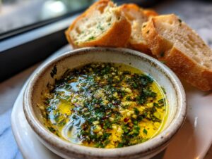 Close-up of a rustic bowl filled with flavorful olive oil bread dip, topped with garlic and herbs, served with slices of crusty bread.