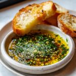 Close-up of a small bowl of seasoned olive oil bread dip topped with fresh herbs and garlic, served with toasted bread slices.