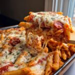 A cheesy, gooey slice of pizza pasta being lifted from a baking dish, showing long cheese pulls.