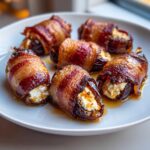 Six glistening, bacon-wrapped stuffed dates filled with creamy cheese, resting on a white plate.