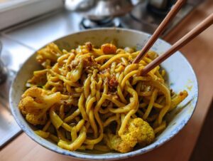 A close-up of a bowl filled with vibrant yellow black pepper turmeric cauliflower and garlic noodles, held by wooden chopsticks.