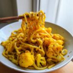 Chopsticks lifting noodles coated in yellow turmeric sauce from a bowl of black pepper turmeric cauliflower and garlic noodles.