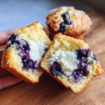 A hand holding a blueberry cream cheese muffin cut in half, showing the rich cream cheese center and blueberries.