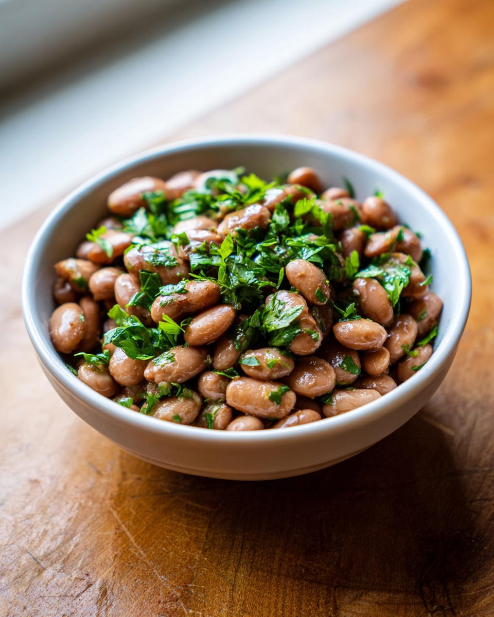 A close-up of a white bowl filled with seasoned healthy pinto beans, topped generously with fresh chopped parsley.