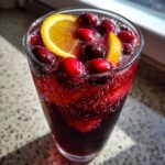 Close-up of a tall glass filled with bubbly cranberry sangria, garnished with fresh cranberries and orange slices.