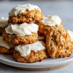A stack of soft, thick carrot cake cookies generously topped with white cream cheese frosting.