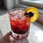 A vibrant, red cherry bourbon smash cocktail filled with crushed ice and garnished with a lemon wheel and two cherries.