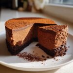 A slice of rich chocolate mousse cake resting next to the main cake on a white plate, dusted heavily with cocoa powder.