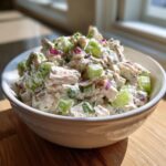 Close-up of a white bowl filled with creamy chicken salad featuring chunks of chicken, bright green celery, and red onion.