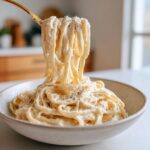 A fork lifts creamy fettuccine coated in rich cottage cheese alfredo pasta sauce, sprinkled with black pepper.