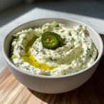 A close-up of creamy jalapeño hummus topped with olive oil and a sliced jalapeño.