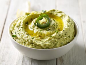 A bowl of vibrant, pale green creamy jalapeño hummus topped with olive oil and a slice of fresh jalapeño.