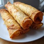 A stack of four golden, crispy cheesy beef flautas, freshly fried and served on a white plate.