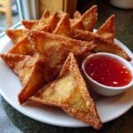 A plate piled high with golden brown, crispy crab rangoons served with a small bowl of bright red sweet chili dipping sauce.