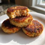 A stack of three golden-brown, crispy salmon cakes on a white plate, with the top one broken open to show the flaky interior.