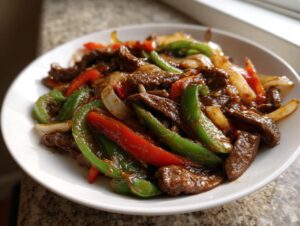 A close-up of a white bowl filled with glossy, saucy pepper steak strips mixed with bright green and red bell peppers and onions.